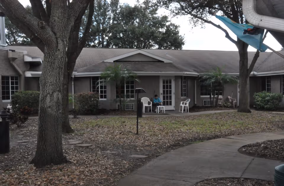 Outdoor view of a single-story building with multiple windows and a door. There are several trees and bushes around the building, a curved concrete pathway, and a person sitting on a white plastic chair near the door. Additional white plastic chairs are also visible nearby.