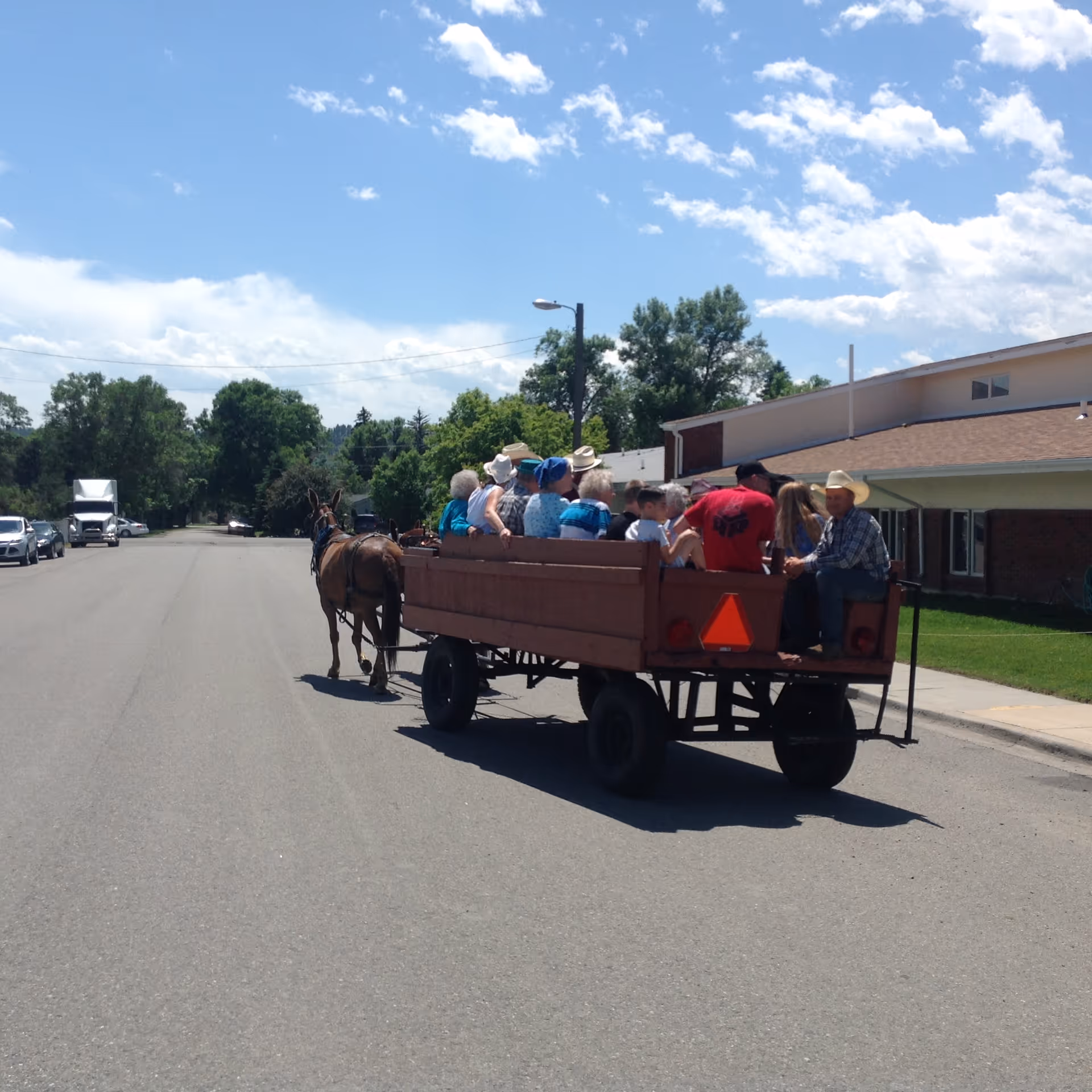 A group of people riding in a wooden wagon pulled by a horse on a paved road near a residential building under a partly cloudy sky.