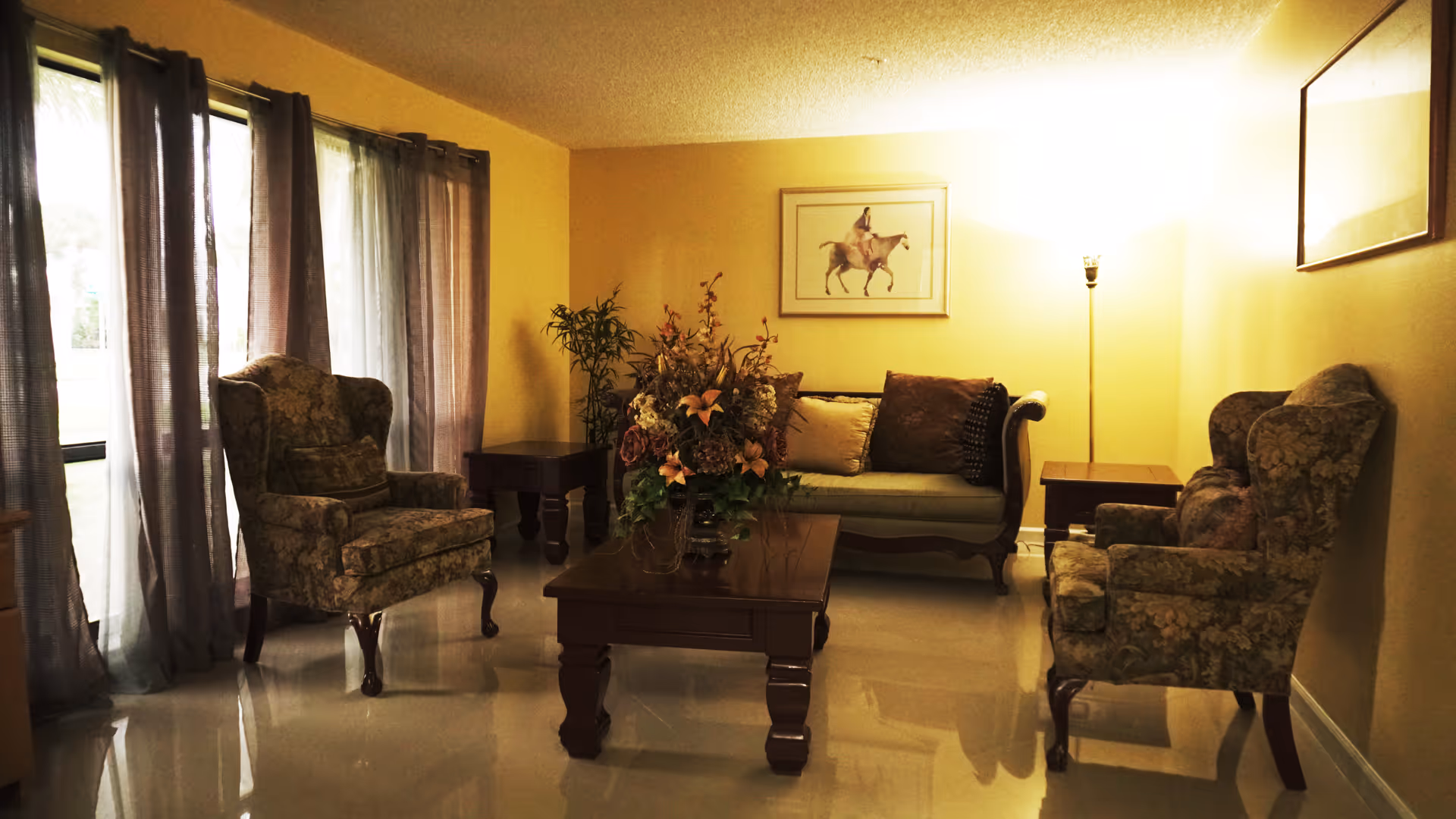A cozy living room with two floral-patterned armchairs, a wooden coffee table with a floral arrangement, a small sofa with cushions, two side tables, a floor lamp, and framed artwork on the walls. Sheer curtains cover large windows letting in natural light.