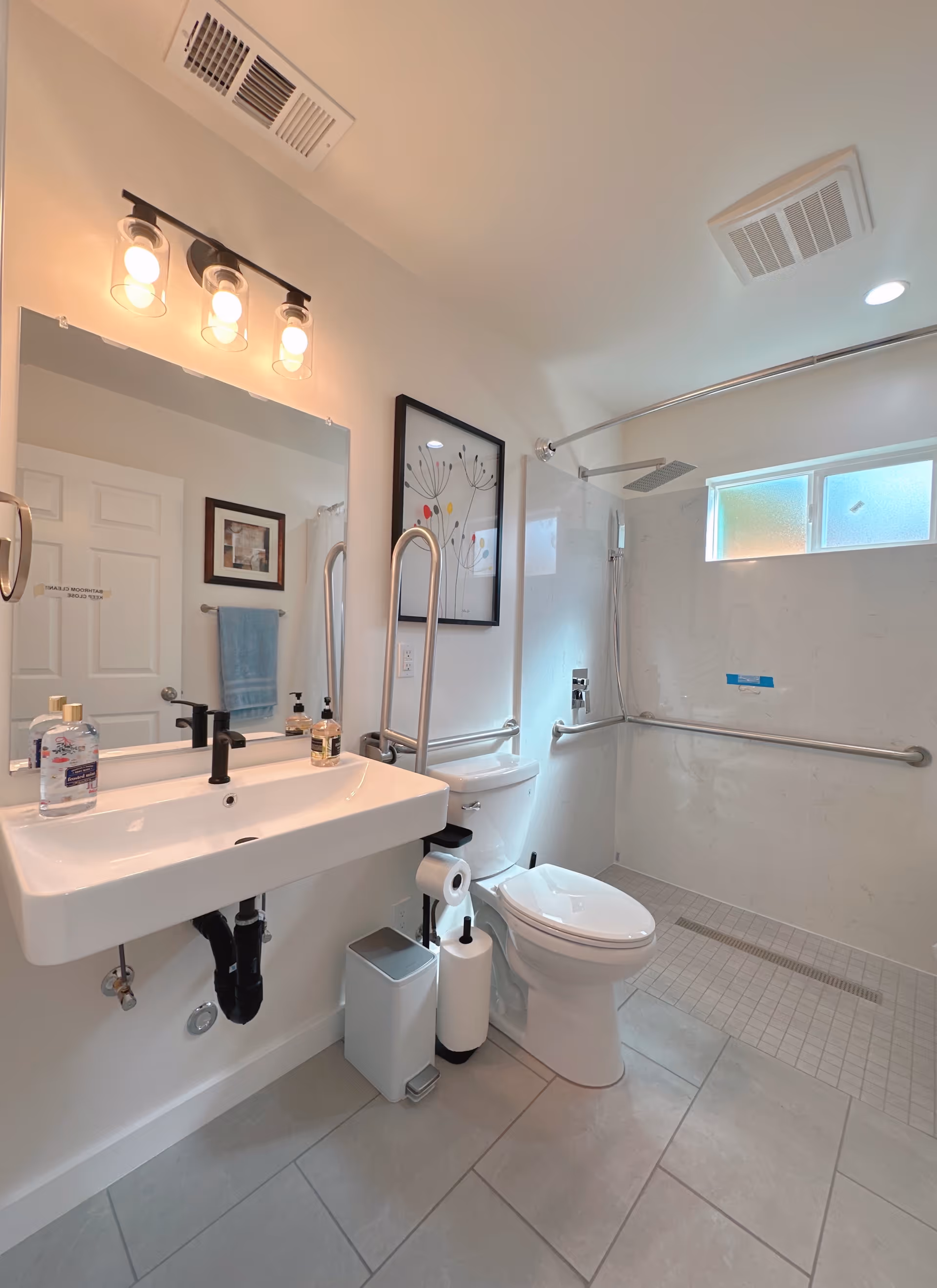 Bright bathroom with sink, mirror, toilet, and tiled shower with grab bars.
