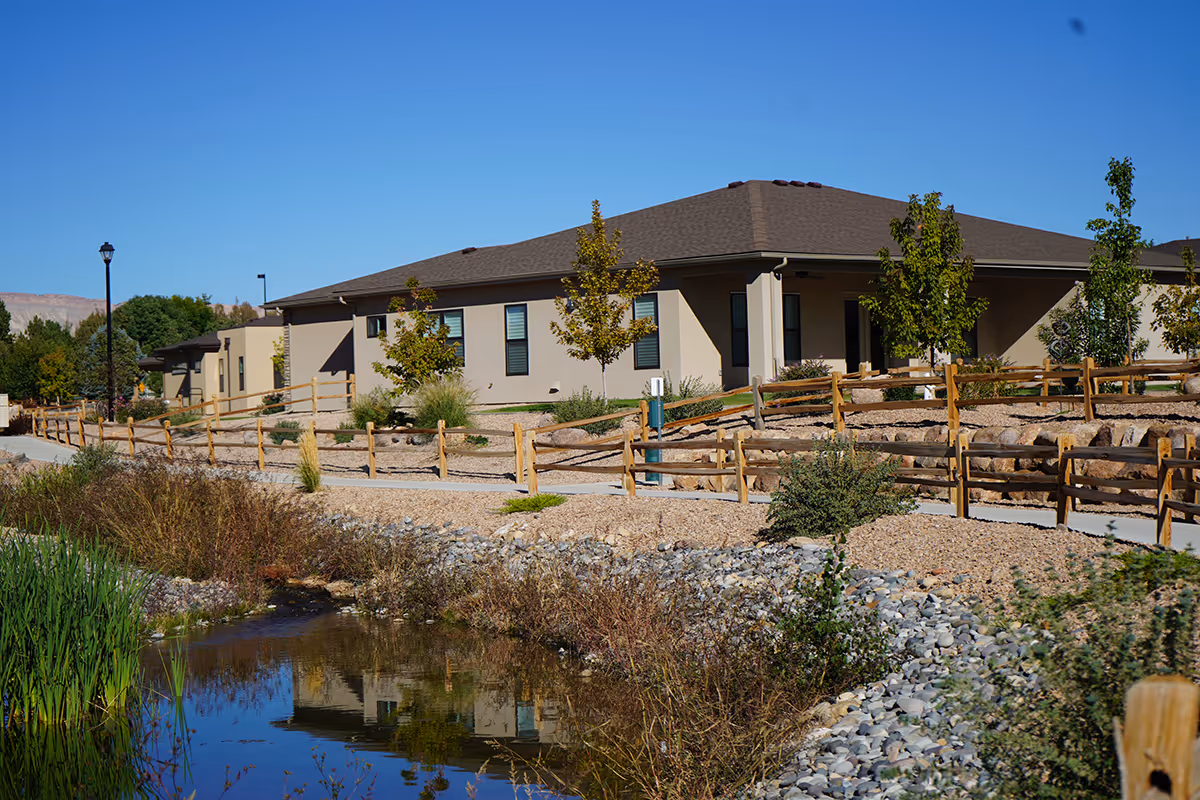 Senior living facility with beige exterior, wooden fences, and pond reflection in foreground.