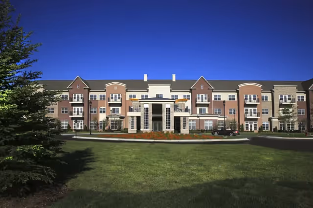 A brick senior living facility with a central glass entrance, balconies, and manicured lawn.