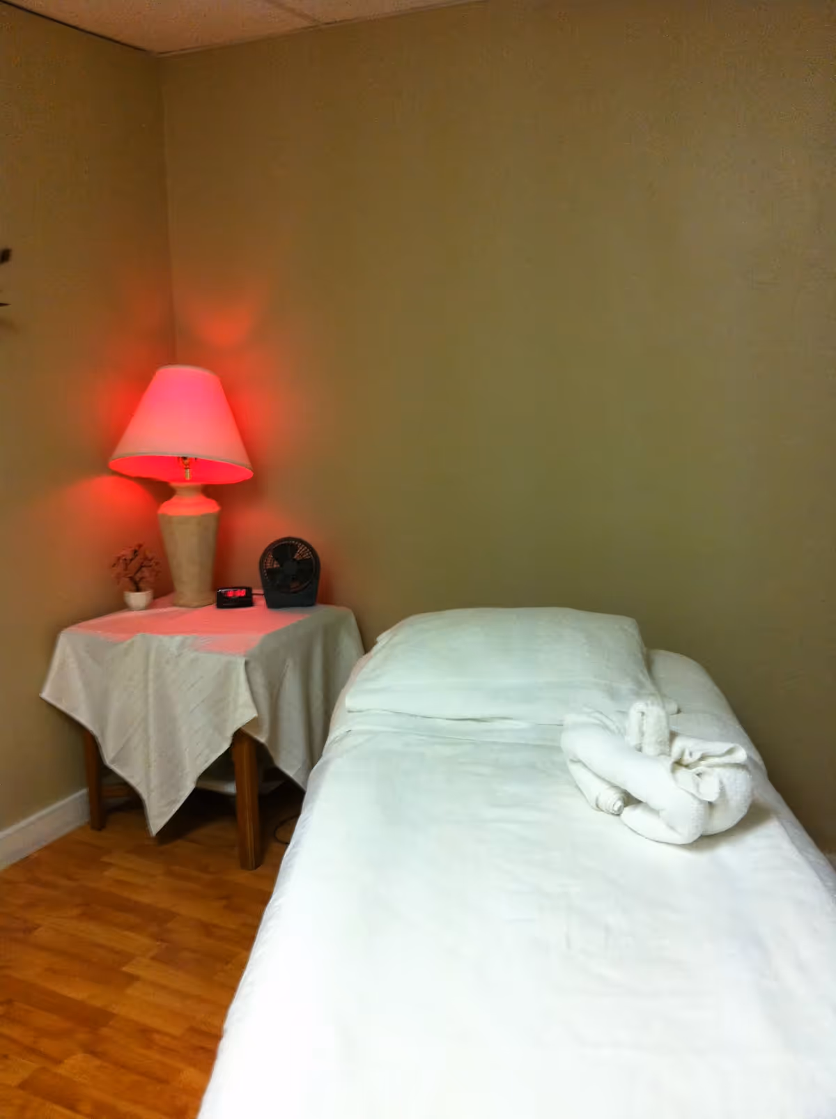 Bedroom scene with single bed, nightstand, pink-lit lamp, folded towel on bed.