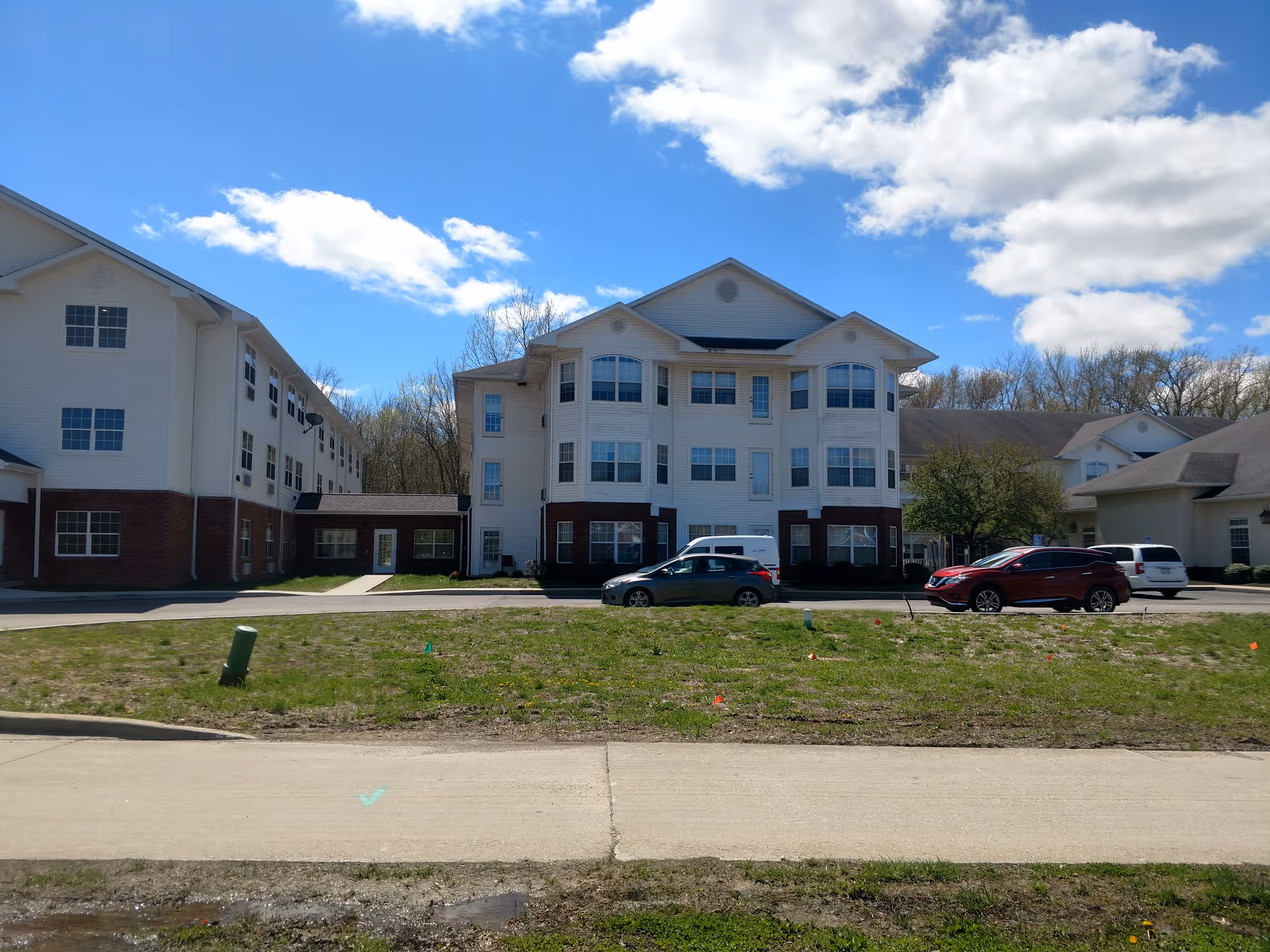 Wide view of a white multi-story senior living building with brick base and cars outside.