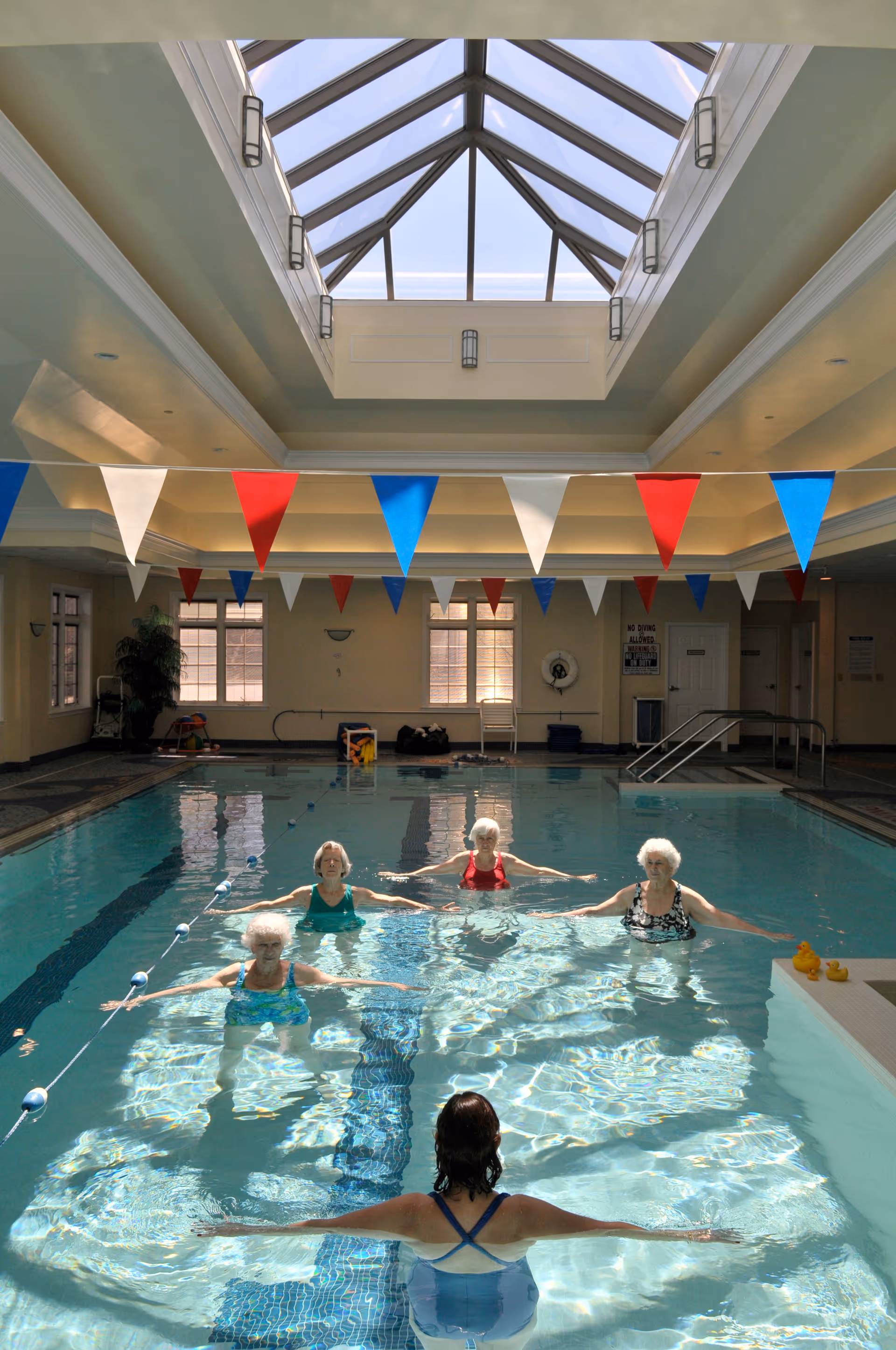 Indoor swimming pool with a glass ceiling and colorful red, white, and blue triangular flags hanging above. Five elderly women are participating in a water exercise class, standing in the pool with their arms extended. The pool area is well-lit with natural light coming through the skylight.