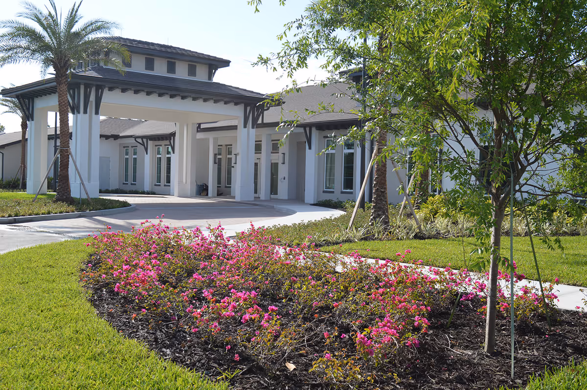Exterior view of Alura Senior Living facility showing the entrance with a covered driveway, surrounded by green grass, palm trees, and a flower bed with pink flowers.