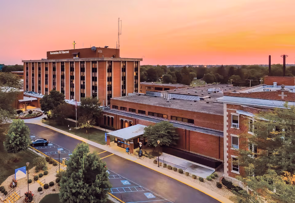 Exterior view of a multi-story brick senior living campus at sunset with entrance and parking.