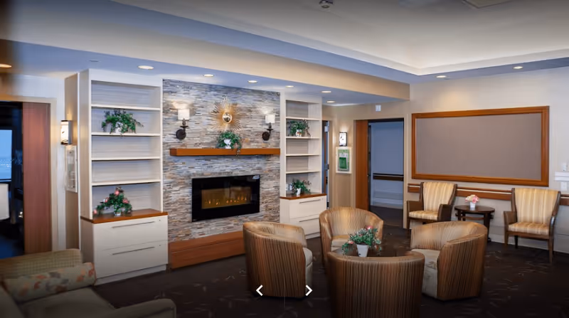 Cozy senior living lounge with a stone fireplace flanked by built-in shelving, a circular arrangement of armchairs, and additional seating along the wall.