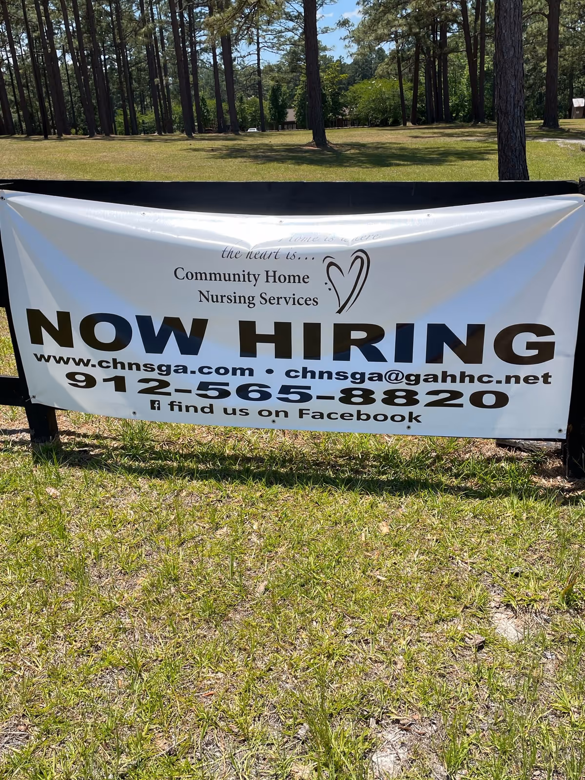 Outdoor banner advertising NOW HIRING for Community Home Nursing Services in a grassy, tree-filled area.