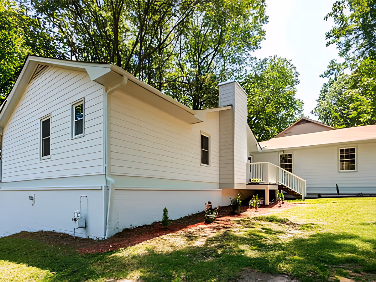 White single-story house with siding, a small porch and stairs, chimney, and a grassy yard with trees behind it.