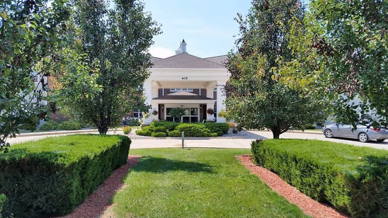 Front exterior view of a senior living facility named Brookstone of Aledo, featuring a white entrance with columns, surrounded by green bushes, trees, and a well-maintained lawn under a clear blue sky.