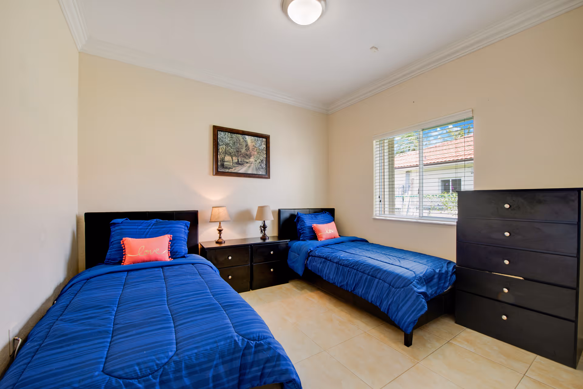 A bedroom with two twin beds featuring blue bedding and red pillows with the word 'love' on them. Between the beds is a black nightstand with two lamps. A framed picture hangs on the beige wall above the nightstand. There is a window with blinds on the right side, and a black chest of drawers is positioned against the right wall. The floor is tiled and the ceiling has crown molding with a ceiling light fixture.