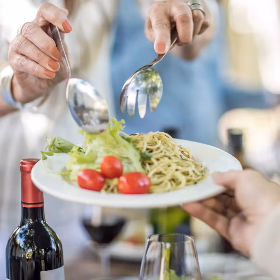 Plate of pasta with tomatoes being served at a dining table, with a wine bottle nearby.