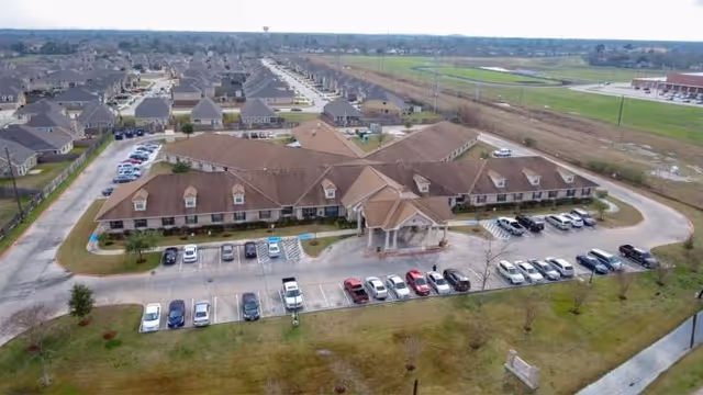 Aerial view of a large single-story building with a brown roof, surrounded by a parking lot with several cars parked. The building is situated in a suburban area with other houses and open fields visible in the background.