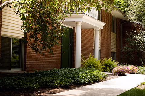 Brick senior living facility entrance with white columns, a green door, and landscaped walkway.