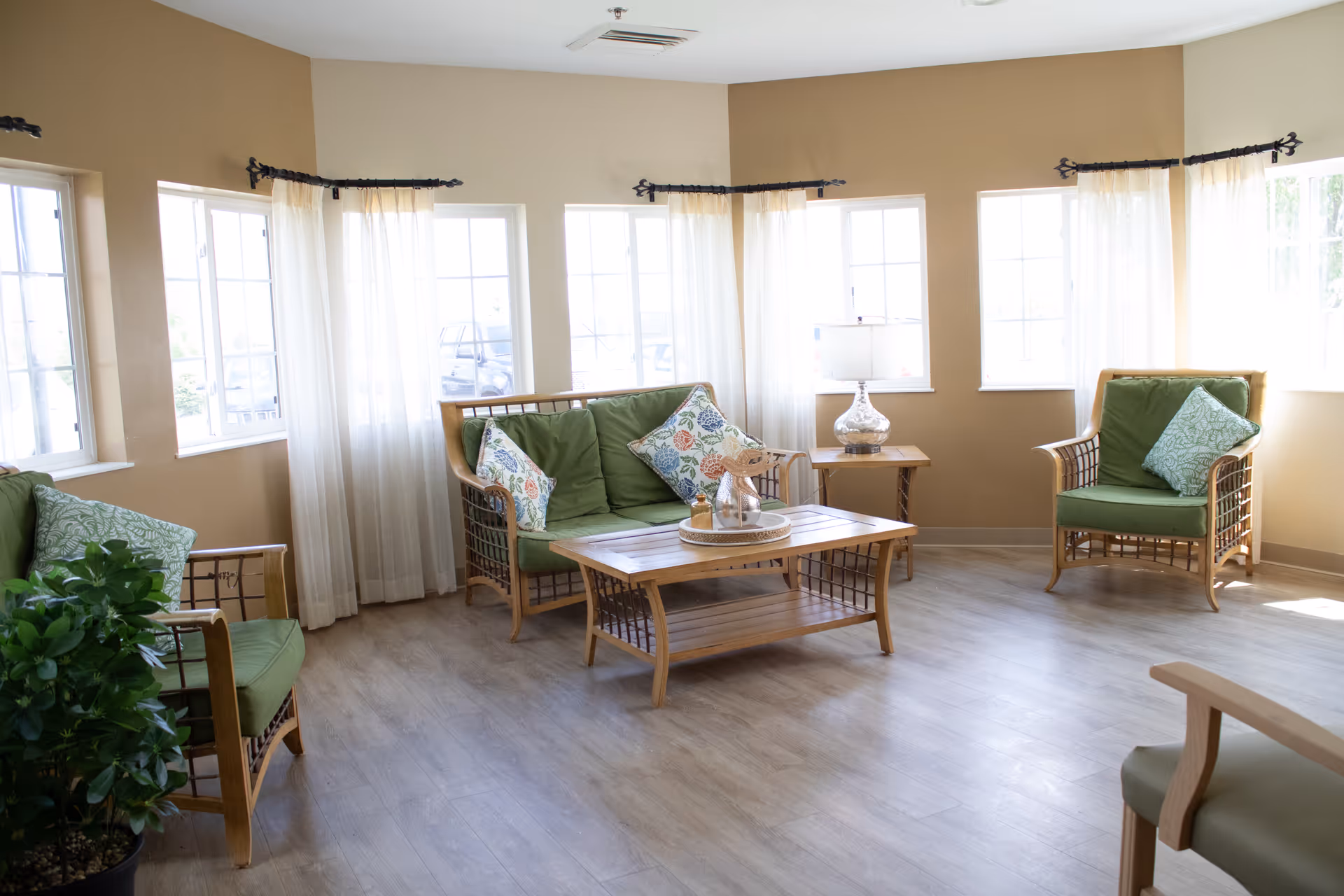 Sunlit senior living facility lounge with wicker seating, green cushions, and a central coffee table.