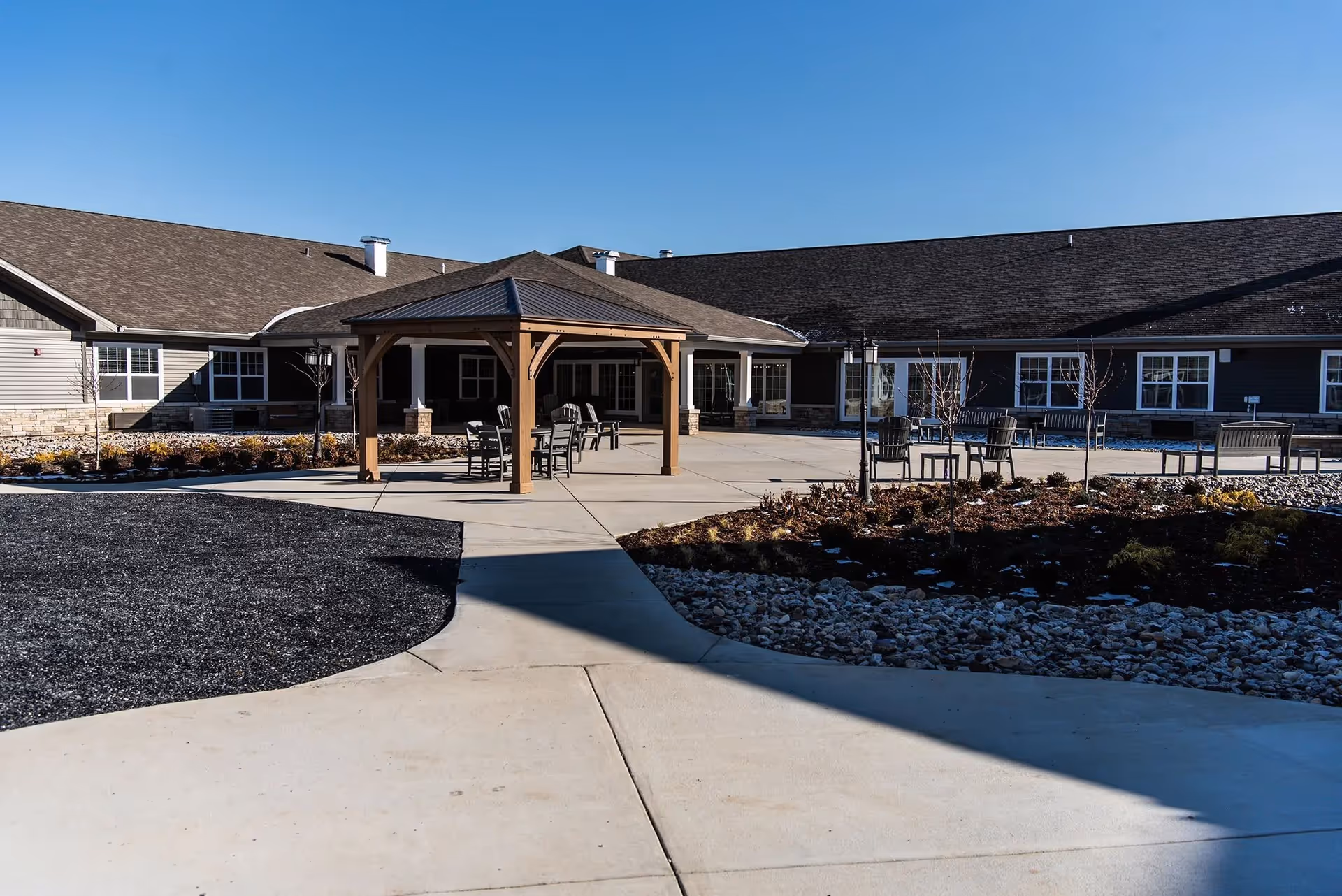 Courtyard with a wooden gazebo, outdoor seating, and a low ranch-style senior living building under a clear blue sky.