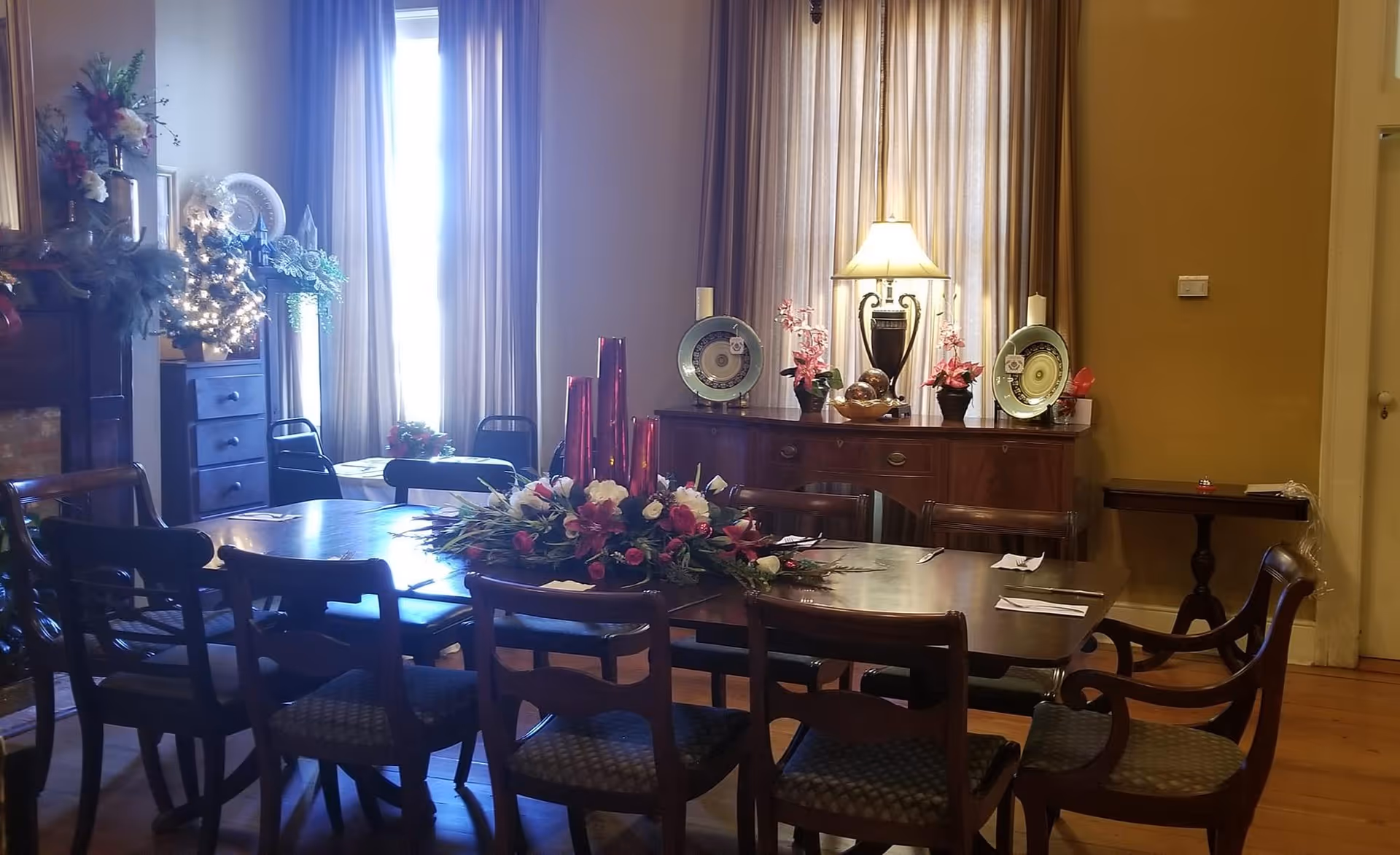 A dining room with a large wooden table surrounded by chairs. The table is decorated with a floral centerpiece and red vases. In the background, there is a sideboard with decorative plates, a lamp, and flower arrangements. Tall windows with curtains allow natural light to fill the room.
