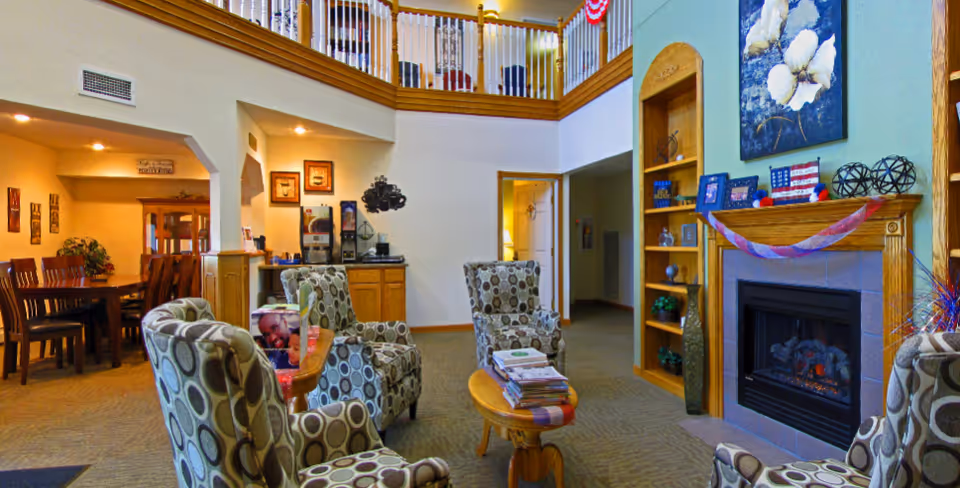 A cozy living room area in a senior living facility with four patterned armchairs arranged around a small wooden coffee table stacked with magazines. There is a fireplace with a wooden mantel decorated with patriotic garlands and framed photos. To the left, there is a dining area with a wooden dining table and chairs. The room has a high ceiling with a balcony railing visible above.