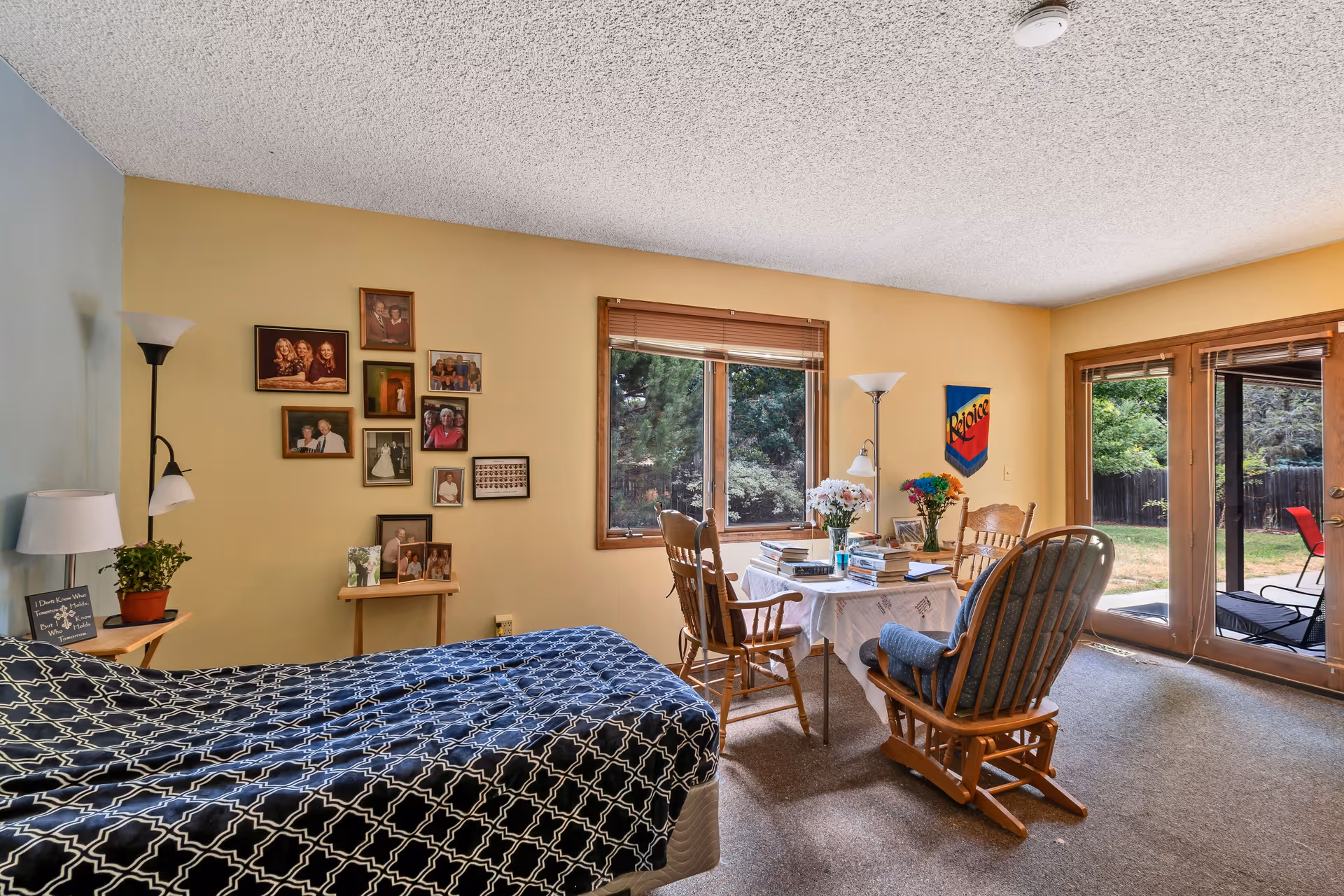 A cozy bedroom with a bed covered in a blue and white patterned blanket on the left side. The walls are painted yellow and light blue, decorated with framed family photos. A small table with a white tablecloth is set with books and flowers, surrounded by wooden chairs. Large windows and glass doors provide a view of a green outdoor garden area.