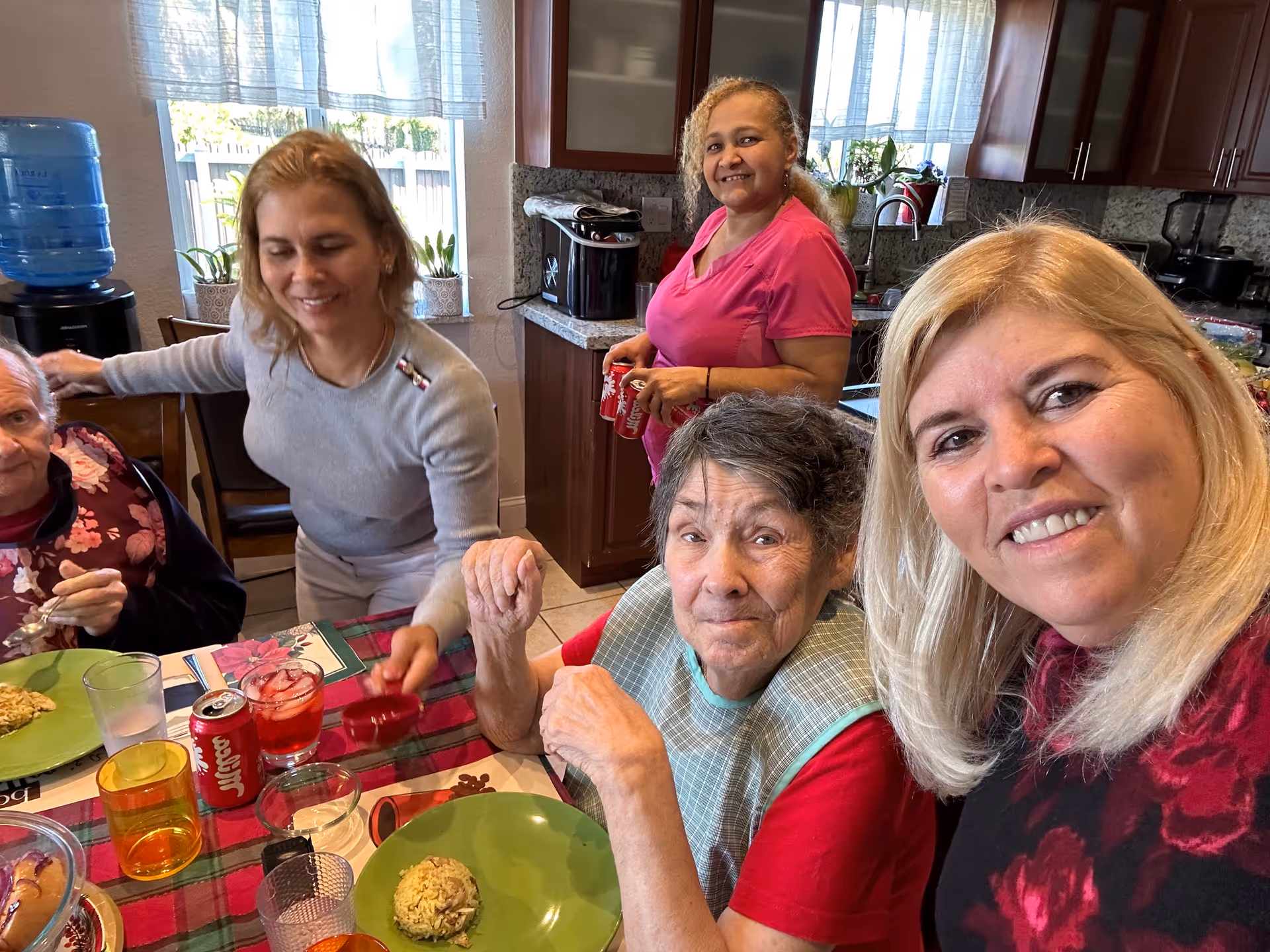A group of five women gathered around a dining table in a kitchen. Two elderly women are seated at the table with plates of food, while three other women stand or lean nearby, smiling. The kitchen has wooden cabinets, a water dispenser, and a window with sheer curtains letting in natural light.