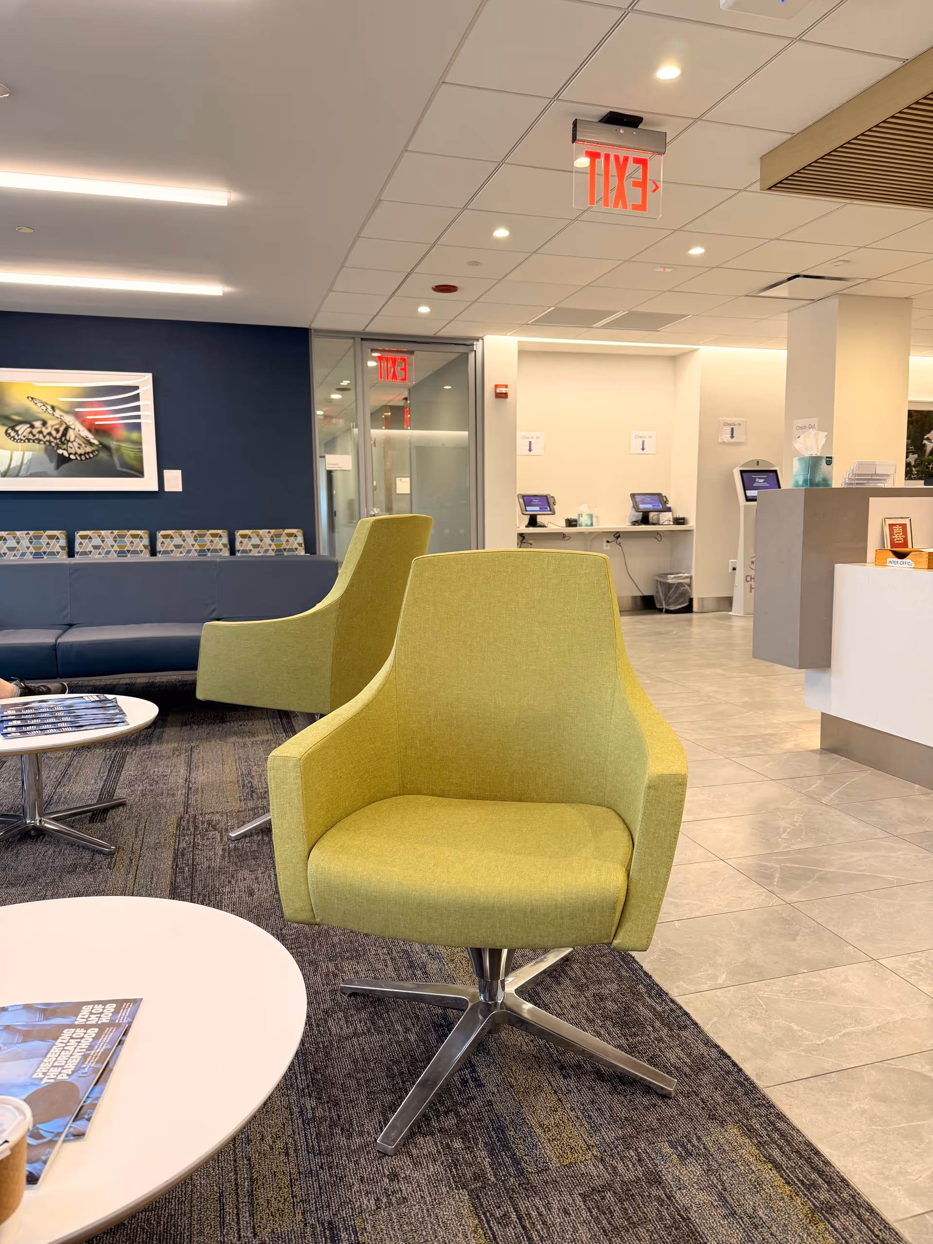 Modern hospital lobby with lime-green chairs, white tables, blue wall art, and a reception desk.