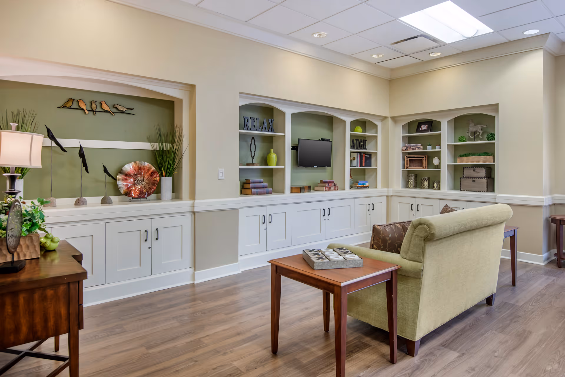Bright lounge area with built-in white cabinets and shelving, a wall-mounted TV, decorative accents, and a green upholstered sofa with side tables.