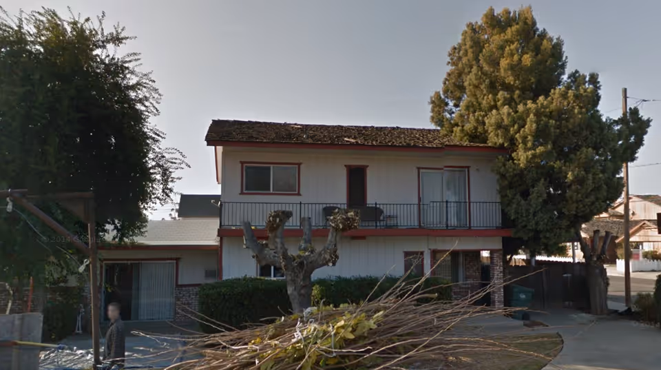 Two-story building with balcony, trimmed trees, and a blurred person nearby.