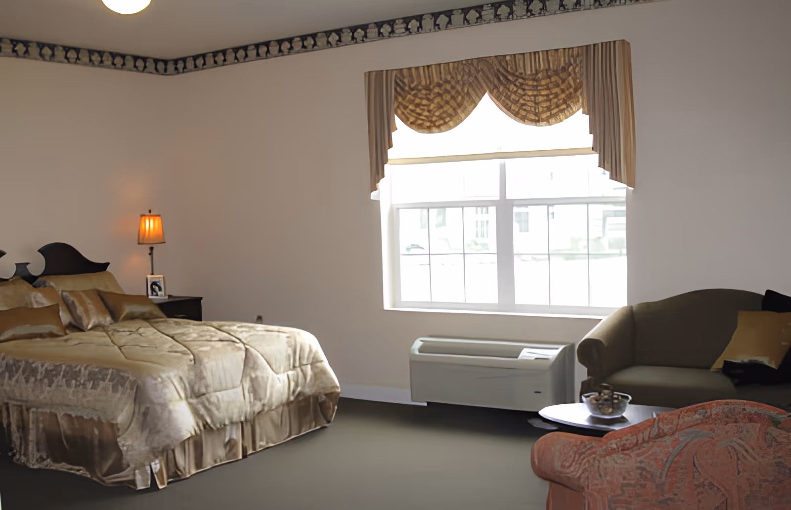 A bedroom in Elmwood Assisted Living of New Bremen featuring a large bed with a beige comforter and pillows, a nightstand with a lamp and a framed photo, a window with patterned valance curtains, an air conditioning unit below the window, and a seating area with a green loveseat and a red patterned armchair around a small round table.