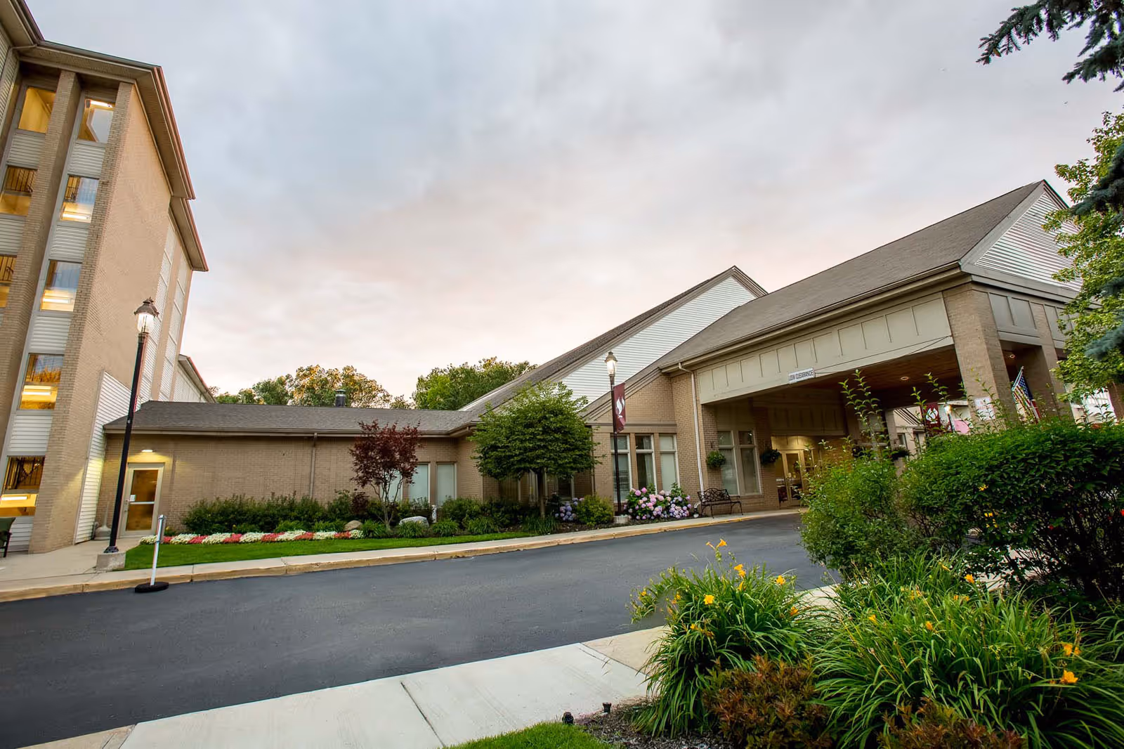 Front entrance and driveway of a multi-story brick senior living building with a covered porte-cochere and landscaped grounds.