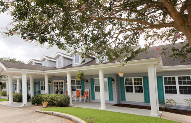 Front entrance of a single-story senior living building with a covered porch, white columns, turquoise shutters, and rocking chairs.