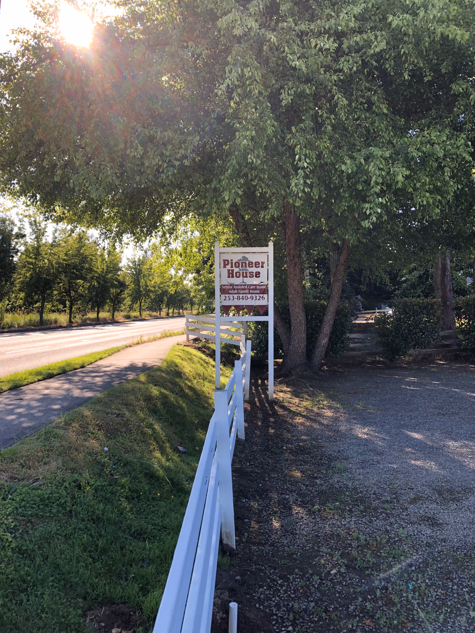 Pioneer House sign beside road under trees with white fence and gravel area.