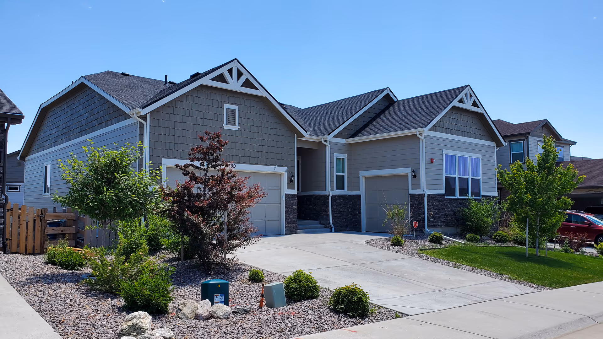 Exterior view of a modern single-story residential building with two garages, a concrete driveway, and landscaped front yard featuring small trees and shrubs under a clear blue sky.