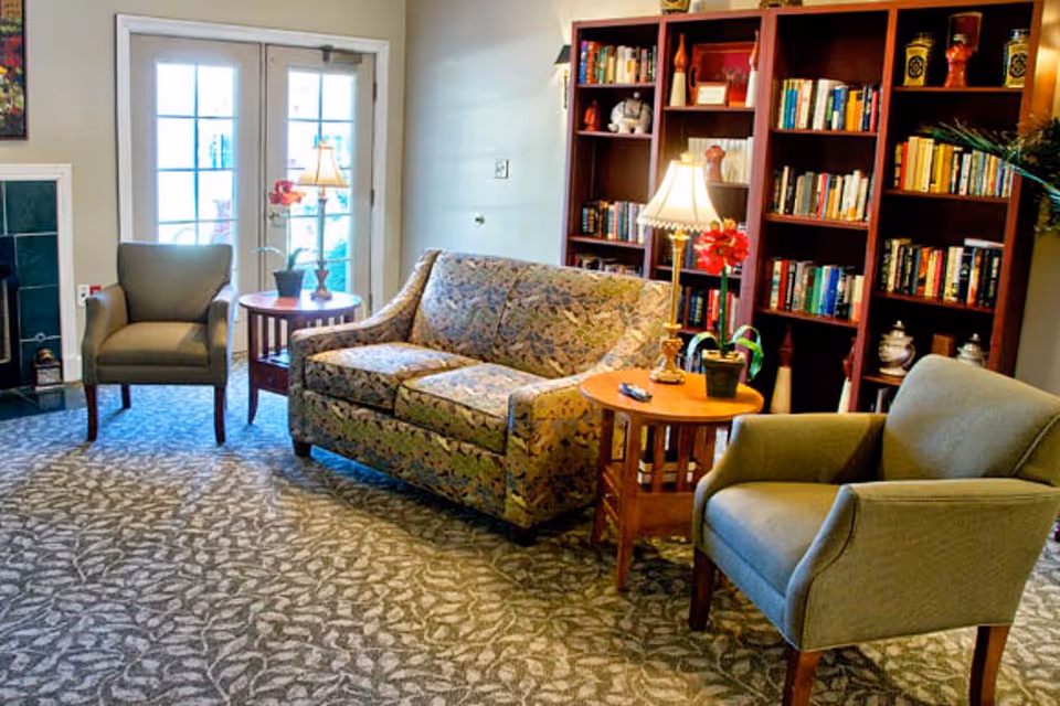 Cozy living room with sofa, two chairs, lamps, and a tall bookshelf beside glass doors.