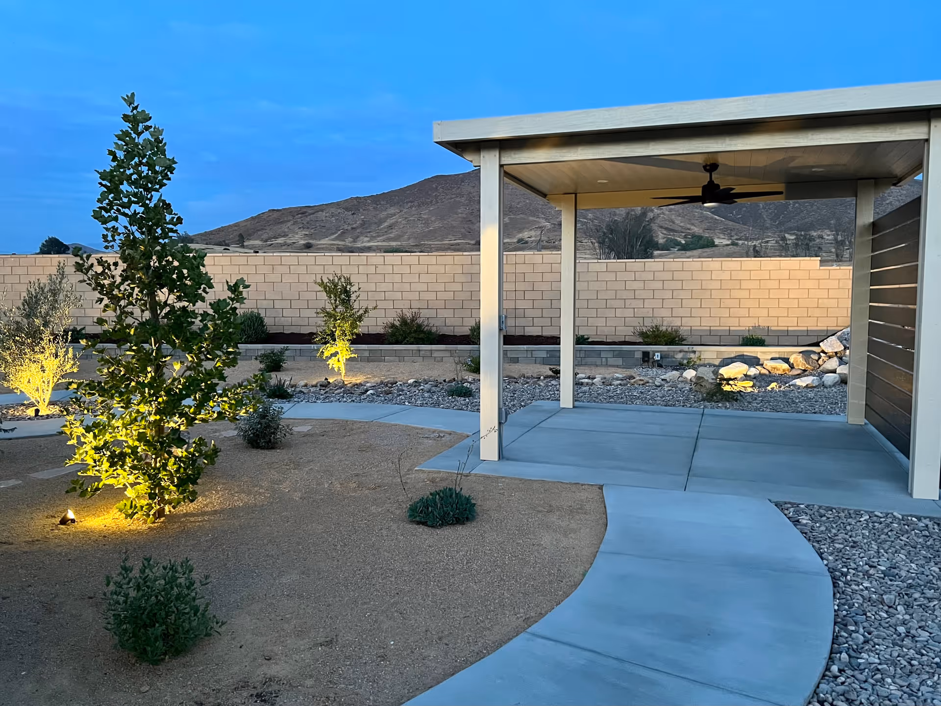 Outdoor patio area with a covered seating space featuring a ceiling fan, surrounded by a landscaped garden with small trees and shrubs illuminated by ground lights, a curved concrete pathway, and a brick wall with hills in the background during dusk.