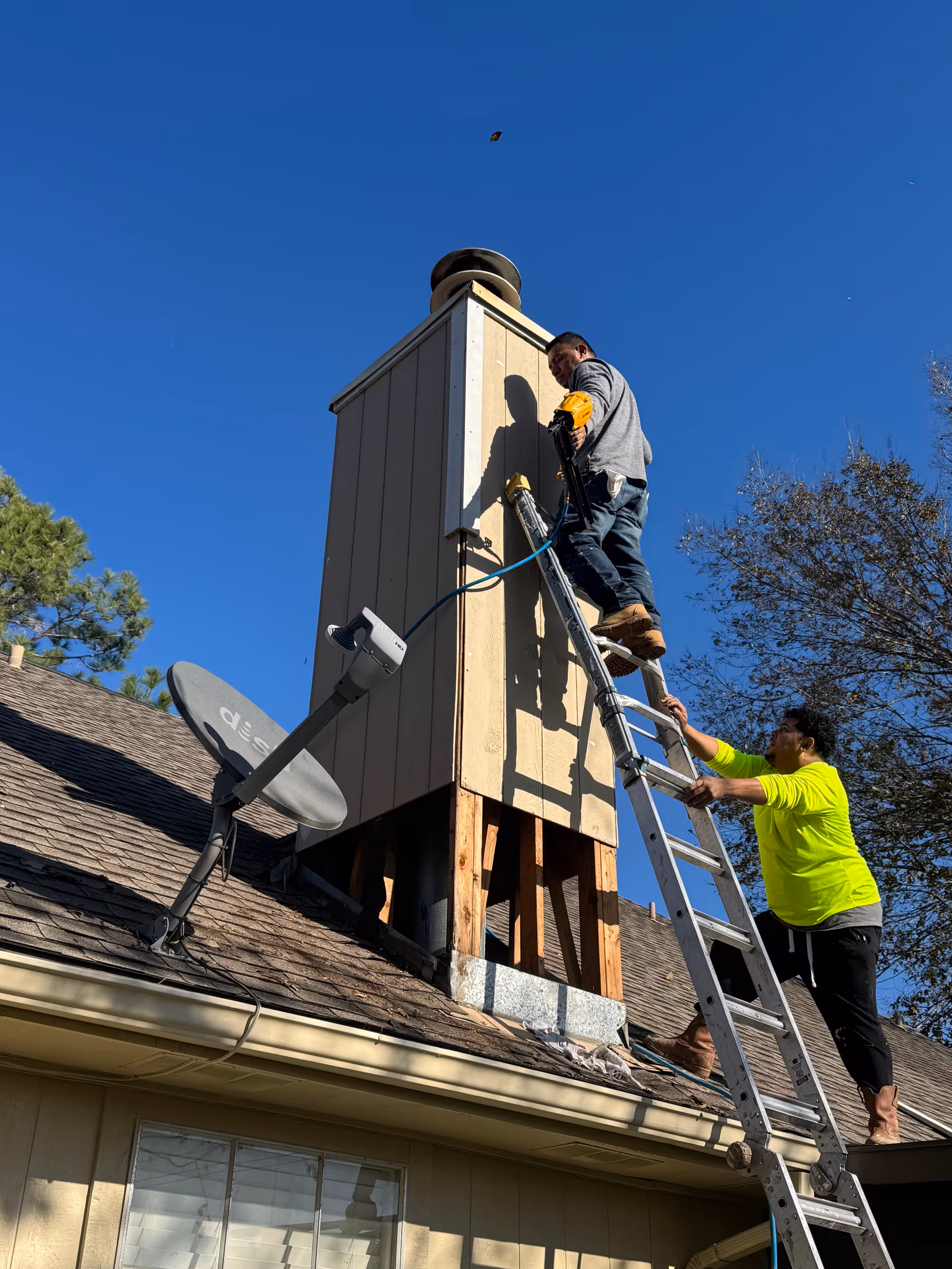Two workers on ladders repair a small structure on a house roof under clear blue sky.
