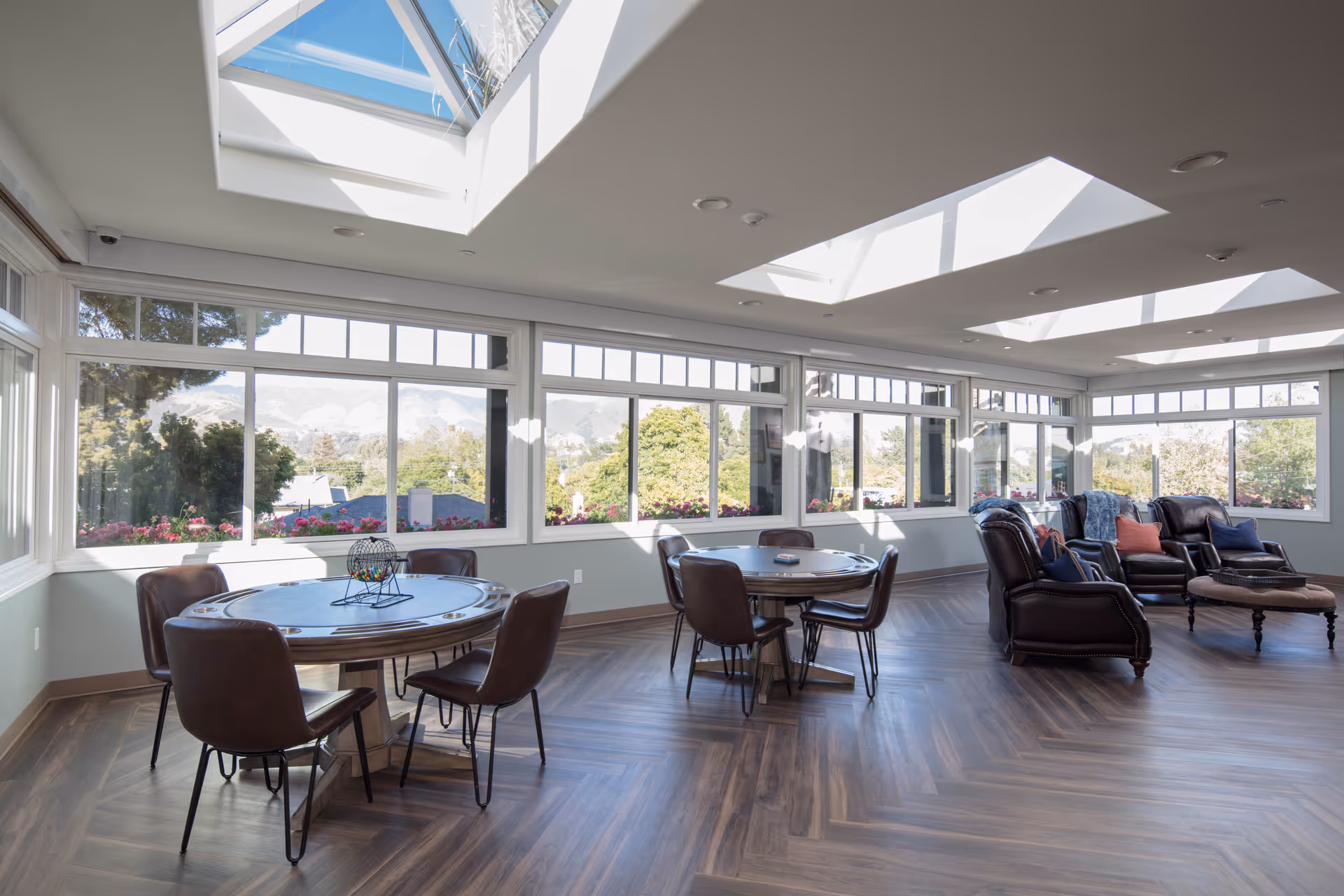 Bright sunlit senior living lounge with skylights, windows, round tables, and leather seating.