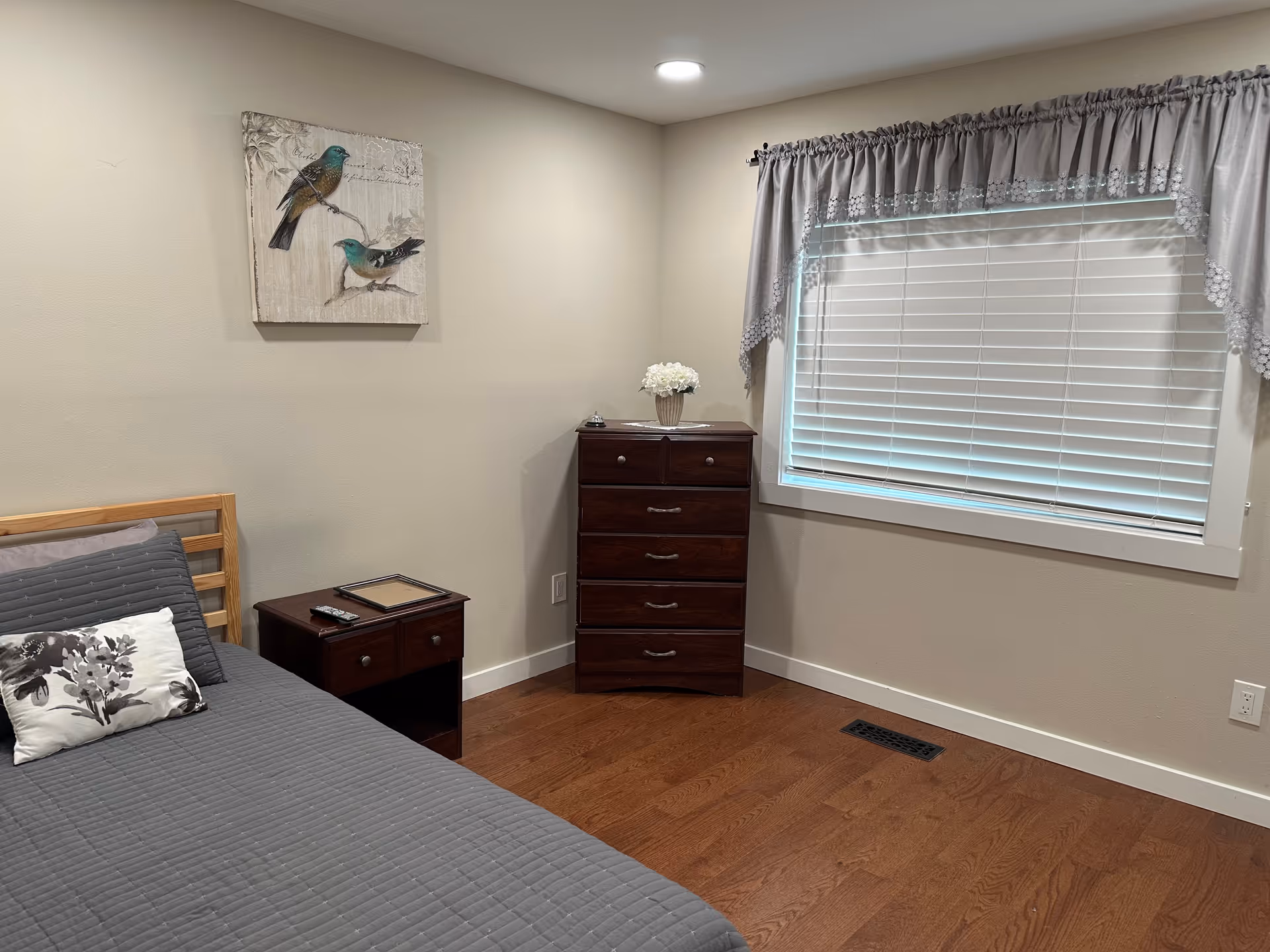A tidy bedroom featuring a bed, dresser, nightstand, and window with blinds.