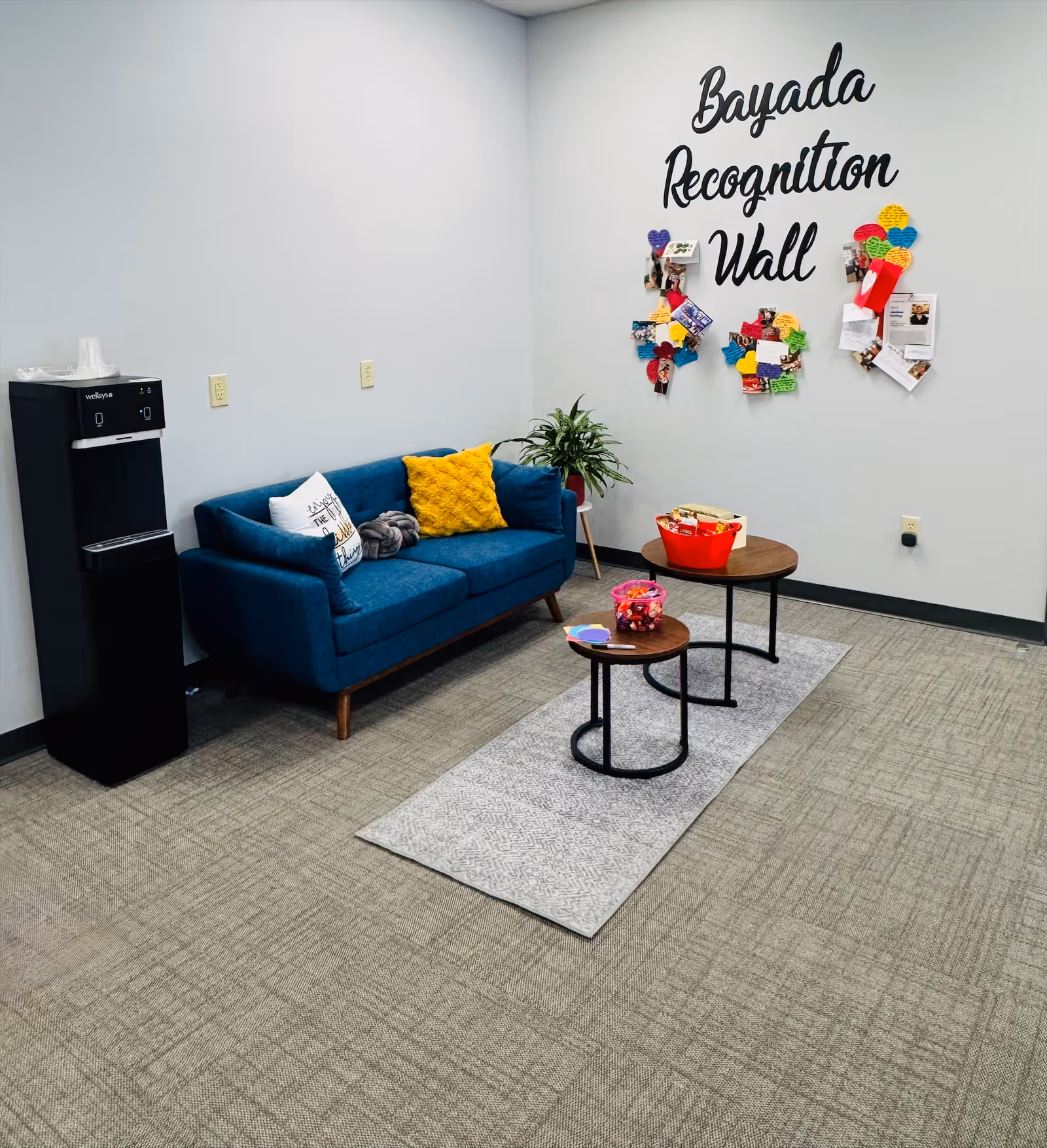 Bright interior lounge with blue sofa, colorful pillows, a Bayada Recognition Wall, and small tables.