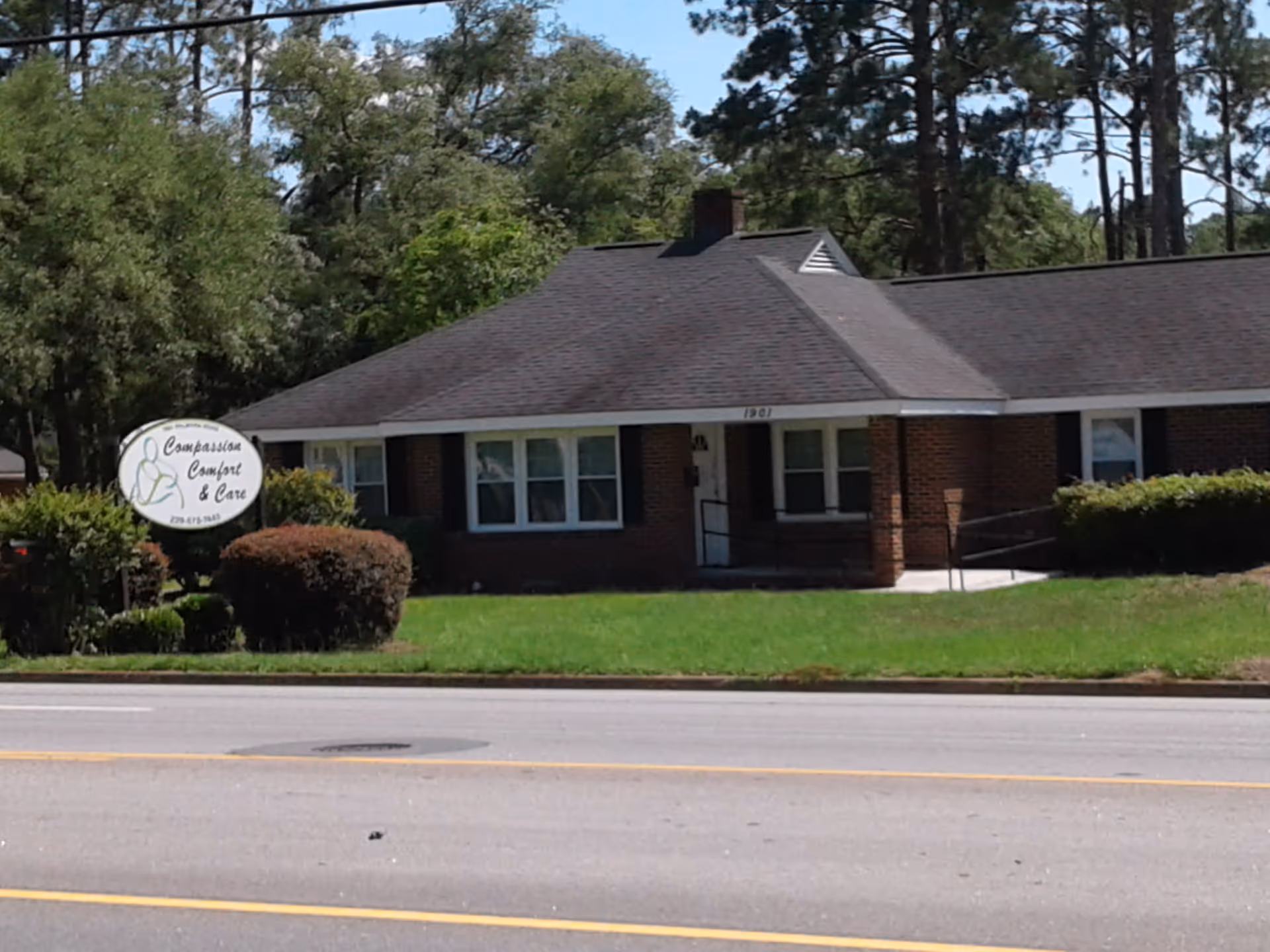 Brick single-story facility with a sign reading Compassion, Comfort and Care, front entrance, trees nearby.