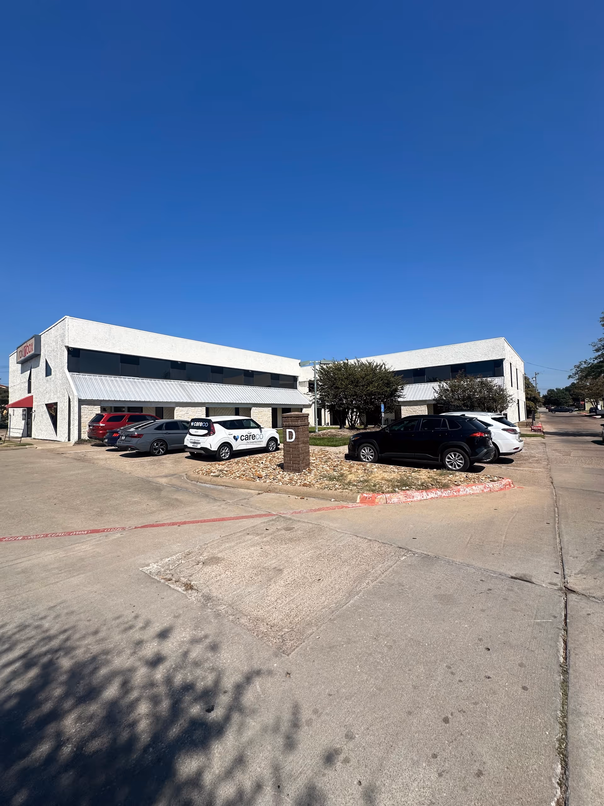 White two-story building with cars parked in a lot under a clear blue sky.