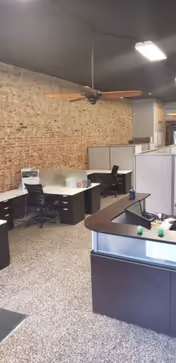 Open interior office area with cubicles, desks, black chairs, and a brick wall.