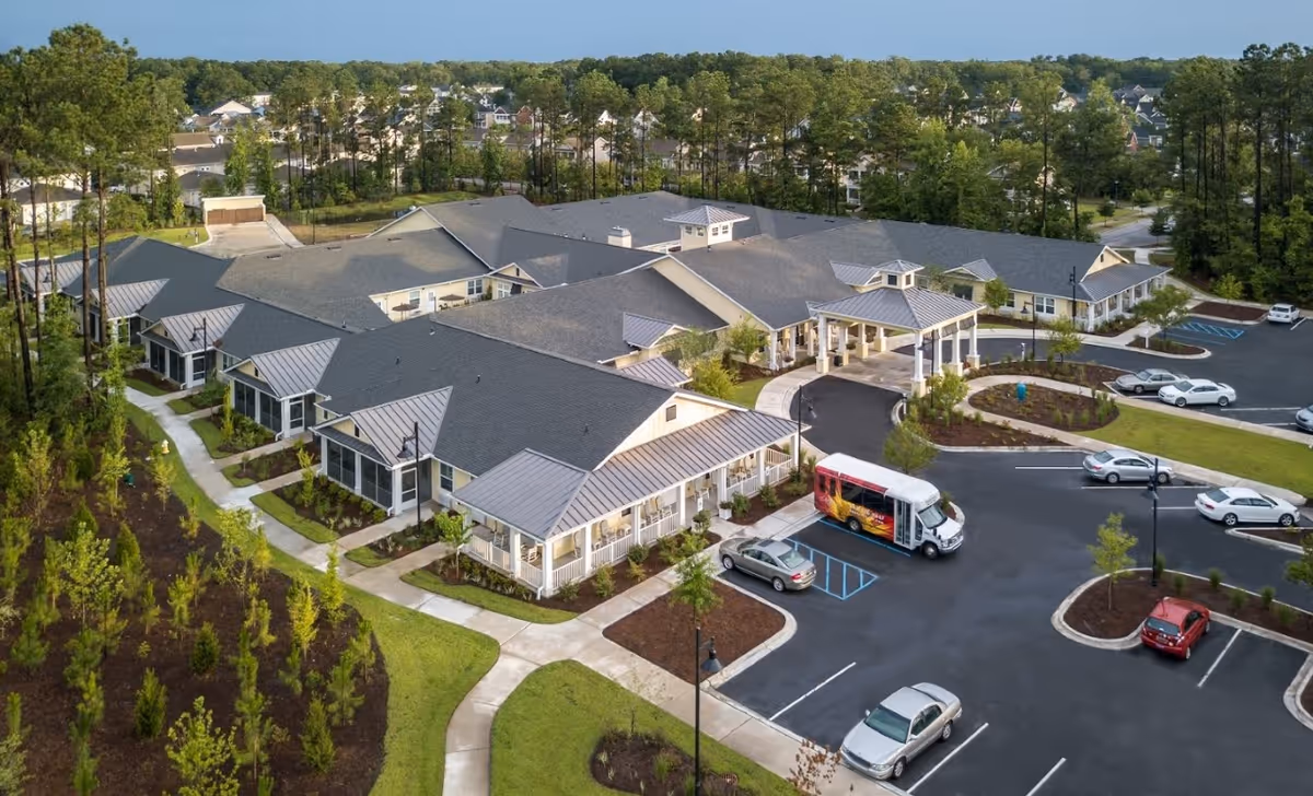 Aerial view of The Addison of West Ashley senior living facility showing a large single-story building with multiple roof sections, surrounded by landscaped greenery and parking lots with several cars and a shuttle bus parked near the entrance.