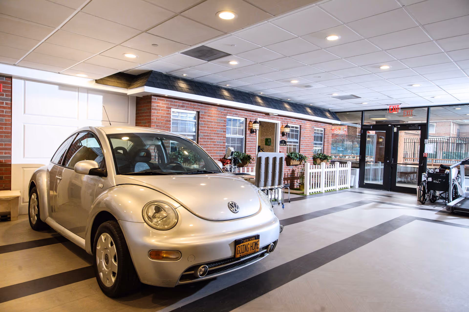 Silver Volkswagen Beetle parked inside a bright senior living facility lobby with brick walls and glass entrance doors.