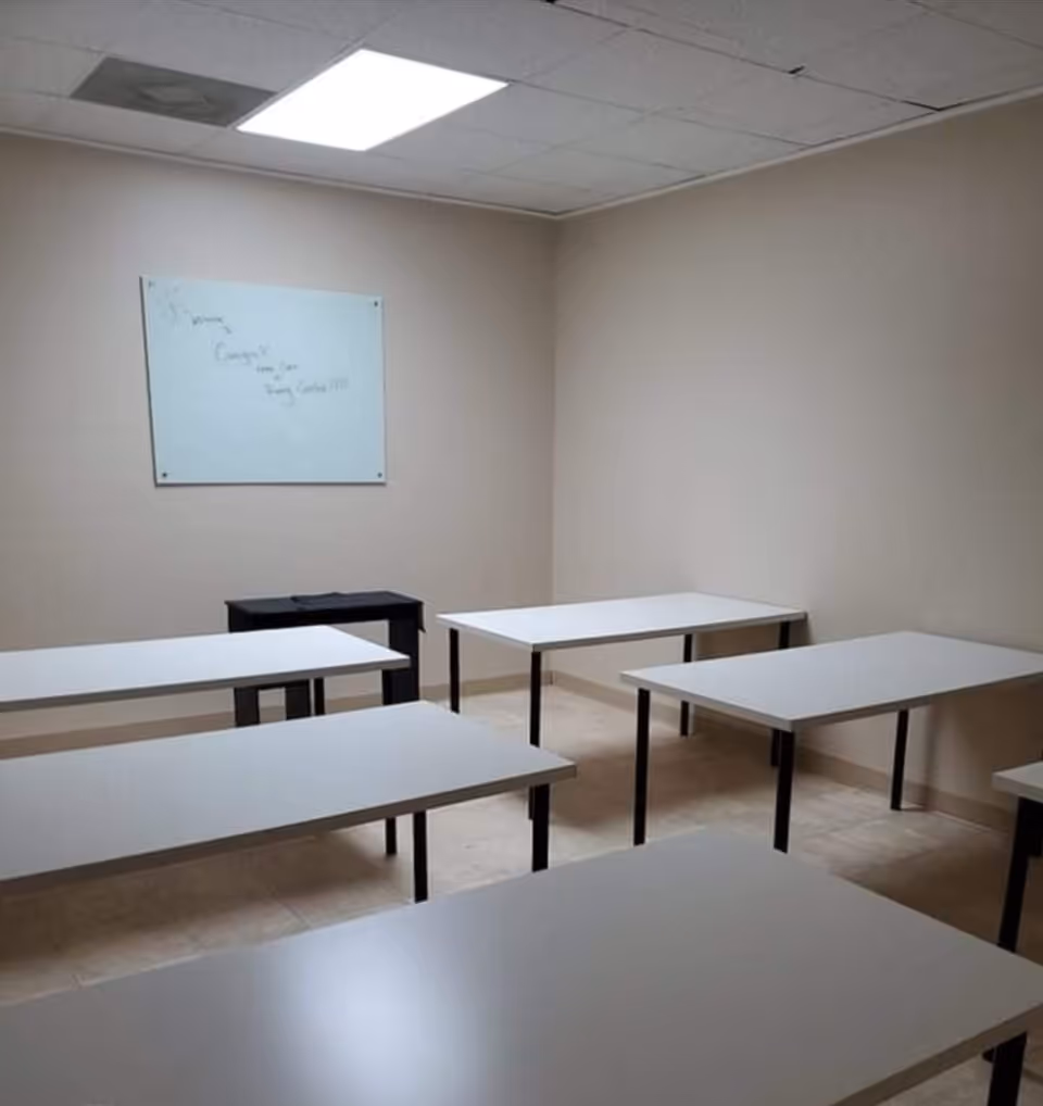 Plain beige interior room with several white desks, a whiteboard, and overhead lighting.