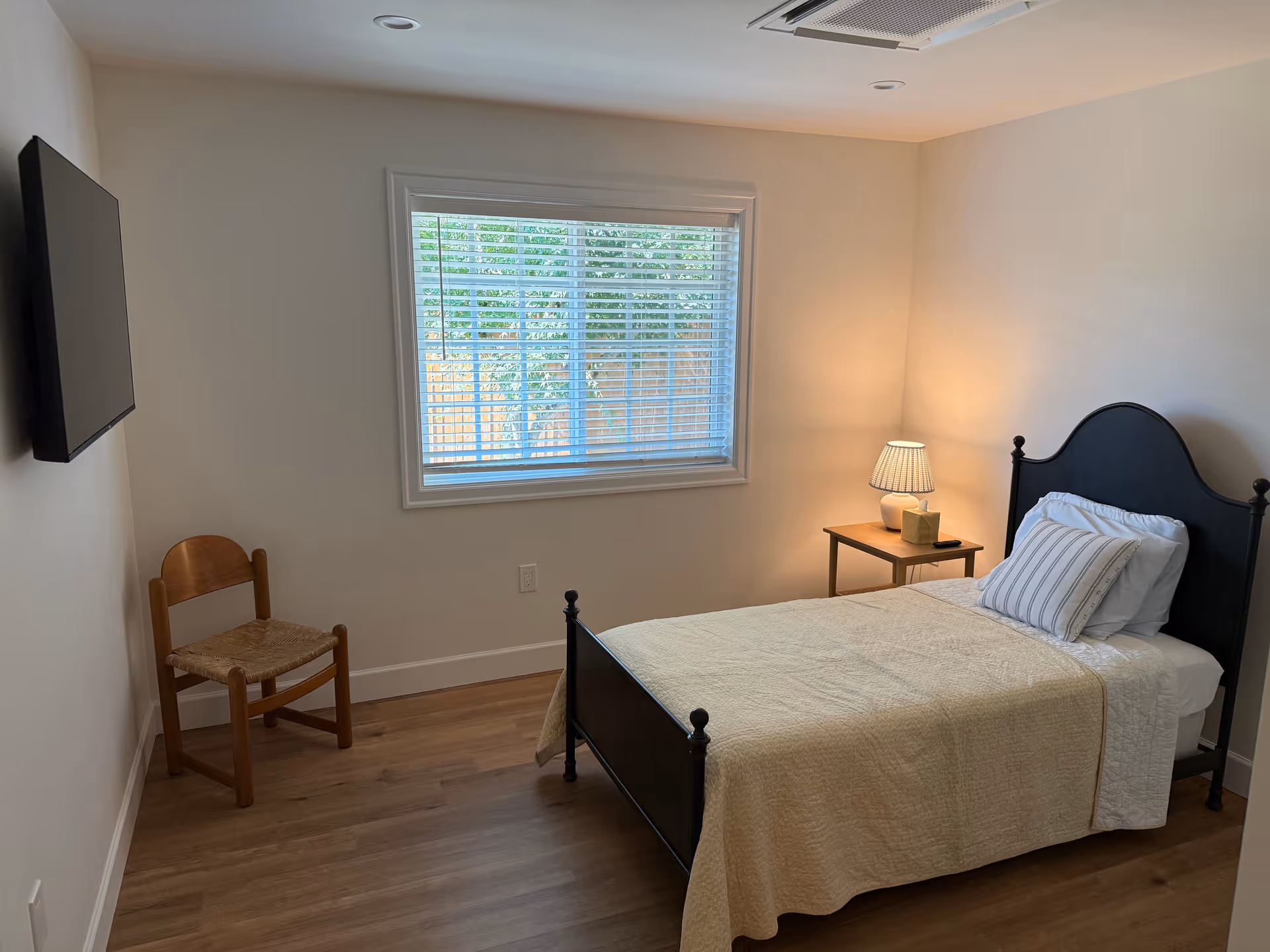 Single bed with headboard, nightstand lamp, chair, and wall-mounted TV in a bright bedroom.