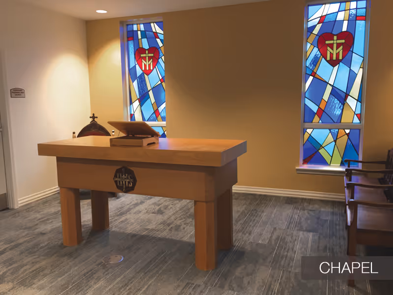 Interior view of a chapel room with a wooden altar table in the center, two stained glass windows with religious symbols on the wall behind it, and wooden chairs along the right side.