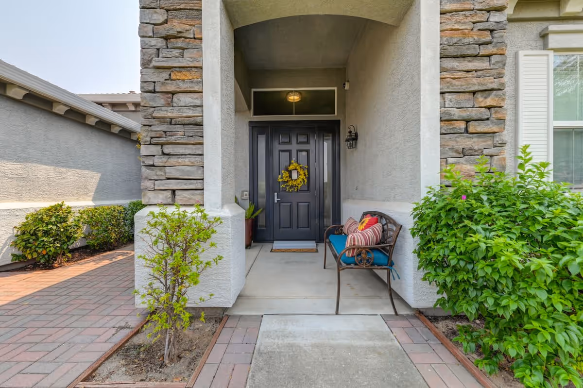 Front entrance of a house with a black door decorated with a yellow wreath, a bench with colorful cushions on the right side, stone and stucco exterior walls, and green shrubs on either side of the walkway.