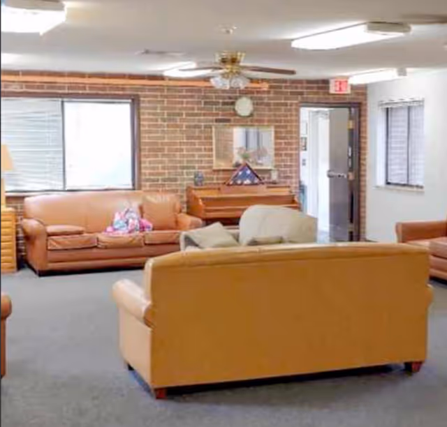 Communal living room with leather sofas and armchairs facing a piano against a brick wall under a ceiling fan.
