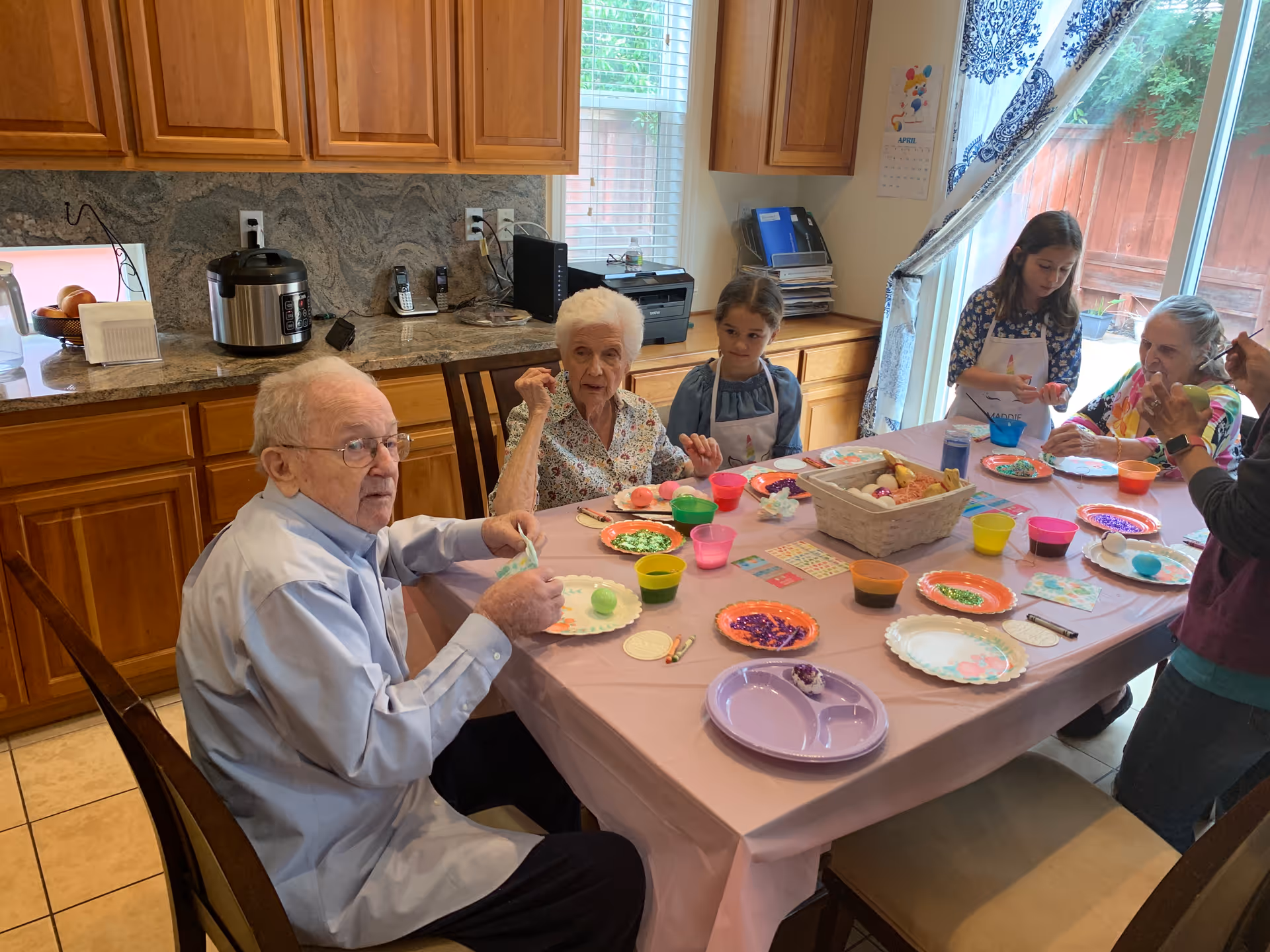 A group of elderly people and children sitting around a table in a kitchen, engaging in an arts and crafts activity with colorful materials and plates on the table. The kitchen has wooden cabinets, a granite countertop, and a window with a view of a wooden fence outside.
